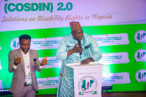 This photo contains the image of the National President of JONAPWD, Abdullahi A Usman, giving his opening remarks as the ongoing COSDIN. He is standing behind a podium on the stage. By his left is a male sign language interpreter