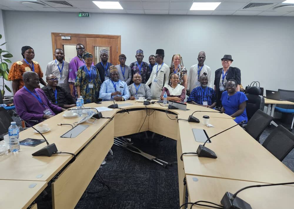 Nineteen participants, men and women, with and without disabilities, gather for a group photo. Seven of them are seated behind a table, representing the leadership of JONAPWD and key members of the World Bank team. Behind them, twelve others stand closely together, forming a cohesive and inclusive backdrop.