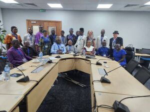 Nineteen participants, men and women, with and without disabilities, gather for a group photo. Seven of them are seated behind a table, representing the leadership of JONAPWD and key members of the World Bank team. Behind them, twelve others stand closely together, forming a cohesive and inclusive backdrop.