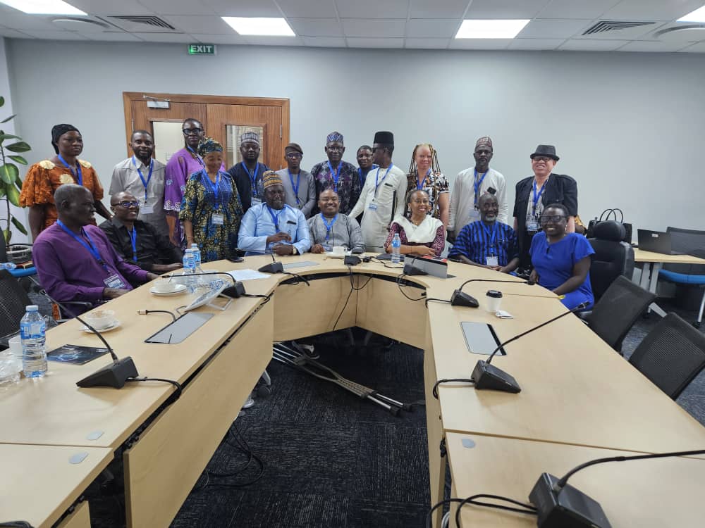 Nineteen participants, men and women, with and without disabilities, gather for a group photo. Seven of them are seated behind a table, representing the leadership of JONAPWD and key members of the World Bank team. Behind them, twelve others stand closely together, forming a cohesive and inclusive backdrop.